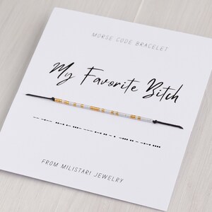 May include: A white card with black text that reads "Morse Code Bracelet" and "My Favorite Bitch" with a black string bracelet with silver and gold beads. The bracelet is in Morse code and the card says "From Miliatari Jewelry".