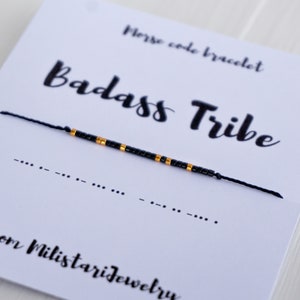 Peut inclure: Un bracelet de perles noires avec des détails dorés, épelant "Badass Tribe" en code Morse. Le bracelet est présenté sur une carte blanche avec le texte "Morse code bracelet" et "from MilitariJewelry".