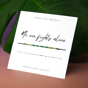 May include: A white card with the text "No one fights alone" and a Morse code bracelet. The bracelet is black with green and gold beads. The card also has Morse code symbols and the text "MORSE CODE BRACELET" and "FROM MILISTARI JEWELRY".