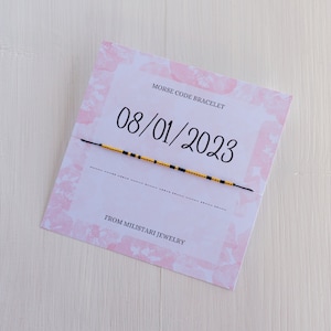 May include: A white card with pink watercolor flowers and the text "MORSE CODE BRACELET" and "FROM MILISTARI JEWELRY". A black string bracelet with gold and black beads is on the card. The date "08/01/2023" is written in black on the card.