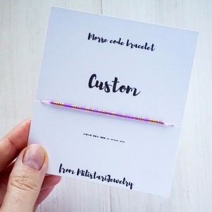 Peut inclure: Un bracelet en perles violettes et dorées avec le mot "Custom" et le code Morse sur une carte blanche. La carte indique également "Bracelet en code Morse" et "de Milistarjewelry".