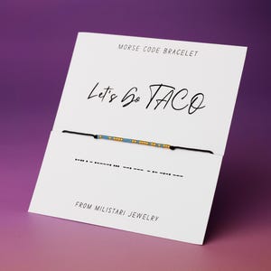 May include: Morse code bracelet on a white card with the text "Let's Go TACO". The bracelet has a black cord with blue and gold beads. The card also includes the text "MORSE CODE BRACELET" and "FROM MILISTARI JEWELRY".