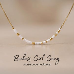 May include: A gold chain necklace featuring white and gold beads. The necklace is curved, with the words "Badass Girl Gang" and "Morse code necklace" in black script below it.