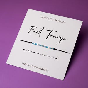 May include: A white card with black text that reads "Morse Code Bracelet" and "Fuck Trump". A black string with blue and silver beads is attached to the card. The string is tied in a knot and the ends are hanging down. The card also has the text "From Miliatari Jewelry".
