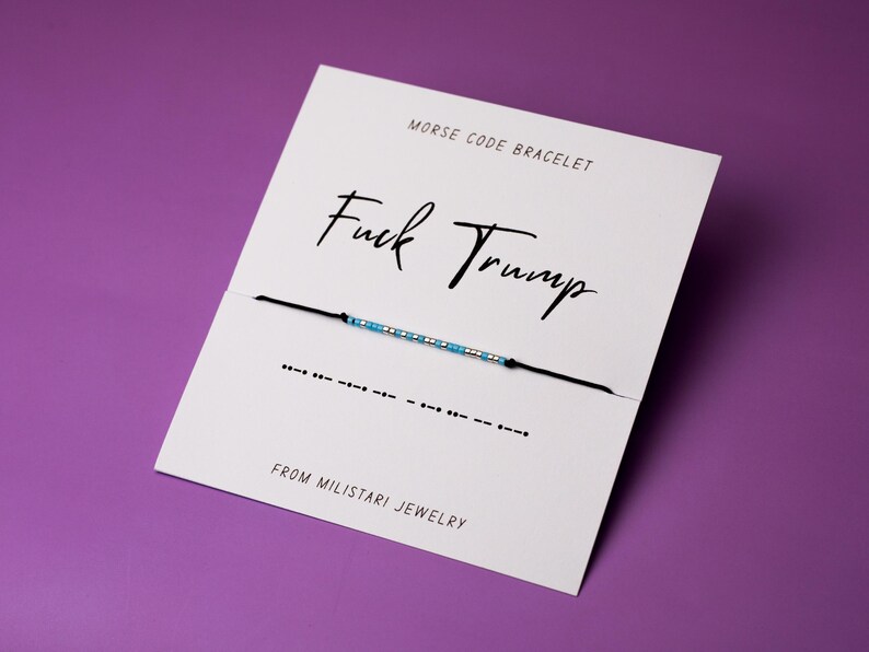 May include: A white card with black text that reads "Morse Code Bracelet" and "Fuck Trump". A black string with blue and silver beads is attached to the card. The string is tied in a knot and the ends are hanging down. The card also has the text "From Miliatari Jewelry".