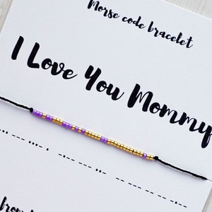 May include: A black string bracelet with purple and gold beads that spells out "I Love You Mommy" in Morse code. The bracelet is on a white card with the text "Morse code bracelet" and "I Love You Mommy" written in black.
