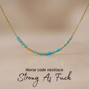 May include: A gold-colored chain necklace with a curved row of turquoise and gold-colored beads. The text "Morse code necklace" and "Strong As Fuck" are below the necklace.