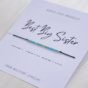 May include: A blue and gold beaded bracelet with a black string. The bracelet is on a white card with the text "Morse Code Bracelet" and "Best Big Sister" in black cursive writing. The card also includes the Morse code for "Best Big Sister" and the text "From Miliatari Jewelry".