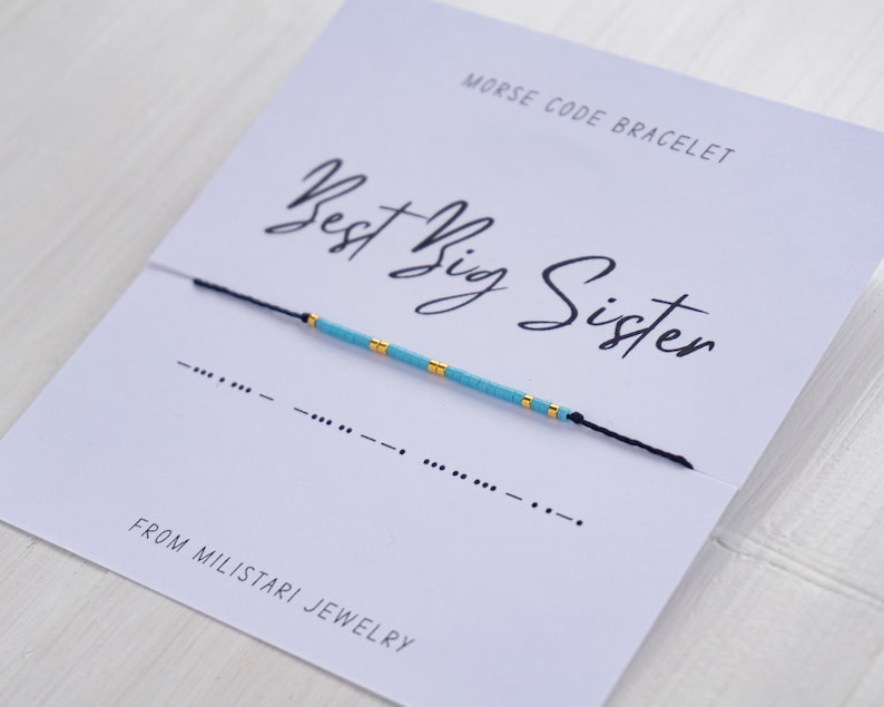 May include: A blue and gold beaded bracelet with a black string. The bracelet is on a white card with the text "Morse Code Bracelet" and "Best Big Sister" in black cursive writing. The card also includes the Morse code for "Best Big Sister" and the text "From Miliatari Jewelry".