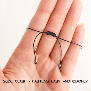 May include: Close-up of a black adjustable cord bracelet with silver beads and a slide clasp. "SLIDE CLASP - FASTENS EASY AND QUICKLY" is printed on the bottom of the image.