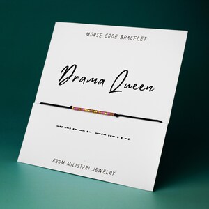 May include: A white card with the text "Drama Queen" in black cursive font. A black string bracelet with pink and gold beads is displayed on the card. The text "MORSE CODE BRACELET" is printed at the top of the card. The text "FROM MILISTARI JEWELRY" is printed at the bottom of the card.