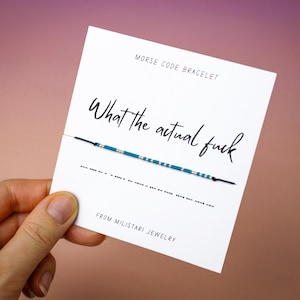 May include: A white card with the text "MORSE CODE BRACELET" and "What the actual fuck" in a handwritten font. Below the text is a bracelet with blue and silver beads. The card is held in a hand against a pink and brown gradient background.