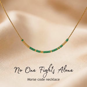 May include: A delicate gold chain necklace featuring a central segment of alternating green and gold cylindrical beads arranged in a Morse code pattern. Below the necklace, the text reads No One Fights Alone and Morse code necklace.