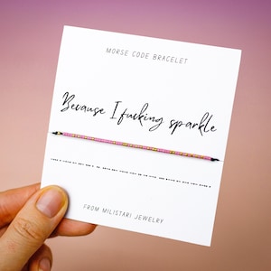 May include: A Morse code bracelet on a white card with the text "Because I fucking sparkle." The bracelet is pink and gold. The card also includes the text "MORSE CODE BRACELET" and "FROM MILISTARI JEWELRY."