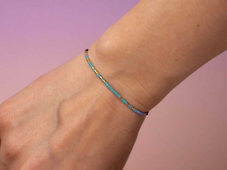 May include: A delicate bracelet made with small turquoise and gold beads. The bracelet is on a person's wrist.