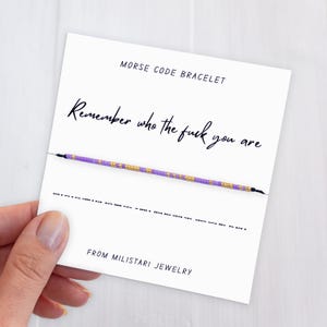 May include: A Morse code bracelet on a white card with the text "Remember who the fuck you are." The bracelet is purple and gold. The card also says "MORSE CODE BRACELET" and "FROM MILISTARI JEWELRY."