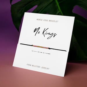 May include: A Morse code bracelet with a black cord and a series of pink and gold beads. The bracelet is displayed on a white card with the words "No Kings" and "From Militari Jewelry".