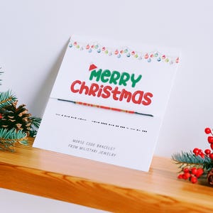 May include: A white card with "Merry Christmas" in green and red, and a Morse code bracelet. The card is decorated with a string of lights and sits on a wooden shelf with pine branches, red berries, and pine cones.