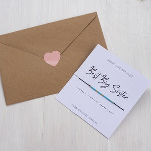 May include: A white card with black text that reads "Best Big Sister" and "Morse Code Bracelet" with a blue and gold string bracelet. The card also says "From Milstani Jewelry".