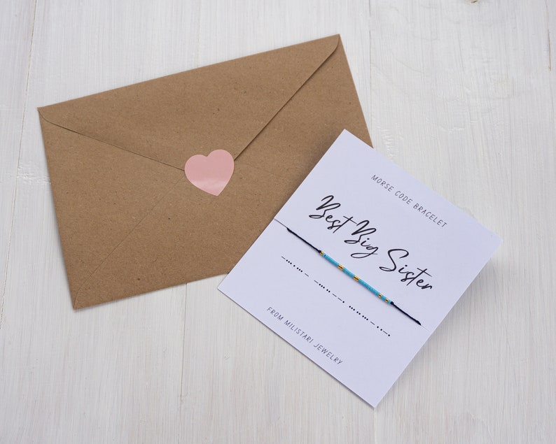 May include: A white card with black text that reads "Best Big Sister" and "Morse Code Bracelet" with a blue and gold string bracelet. The card also says "From Milstani Jewelry".
