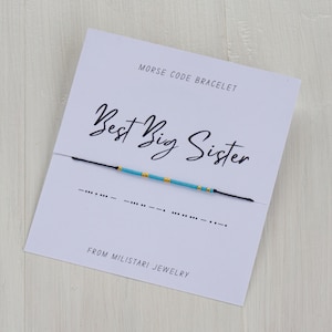 May include: A blue and gold beaded bracelet with the message "Best Big Sister" in Morse code on a white card with the text "Morse Code Bracelet" and "From Miliatari Jewelry".