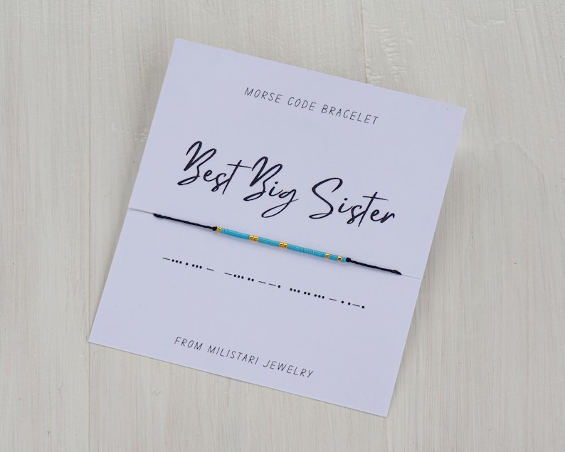 May include: A blue and gold beaded bracelet with the message "Best Big Sister" in Morse code on a white card with the text "Morse Code Bracelet" and "From Miliatari Jewelry".