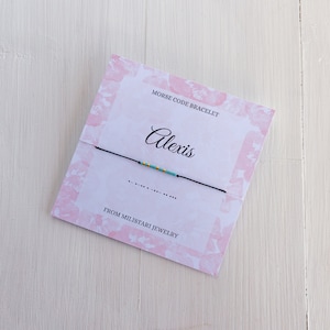 Peut inclure: Un bracelet en cordon noir avec des perles turquoise et dorées qui épellent le nom "Alexis" en code Morse. Le bracelet est sur une carte rose et blanche avec le texte "Bracelet en code Morse" et "De Miliatari Jewelry".