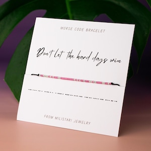 May include: A white card with the text "Don't let the hard days win" in black script. The card also has the text "Morse Code Bracelet" and "From Miliatari Jewelry". A pink and silver beaded bracelet is displayed on the card.