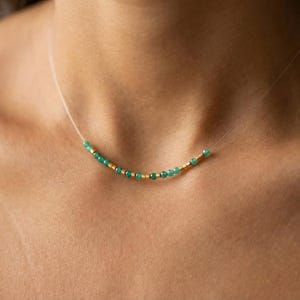 May include: A delicate necklace featuring small, round, emerald green beads interspersed with gold-colored beads. The necklace is strung on a thin, nearly invisible cord, creating a floating effect. The necklace is a minimalist jewelry piece.