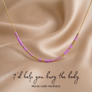 May include: A gold-colored chain necklace with a series of small, lavender and gold beads. The necklace is displayed on a satin fabric. The text "I'd help you bury the body" and "Morse code necklace" is below the necklace.