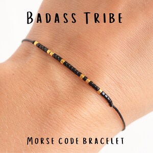 May include: A black string bracelet with gold beads spelling out "BADASS TRIBE" in Morse code. The bracelet is on a person's wrist. Morse code bracelet.