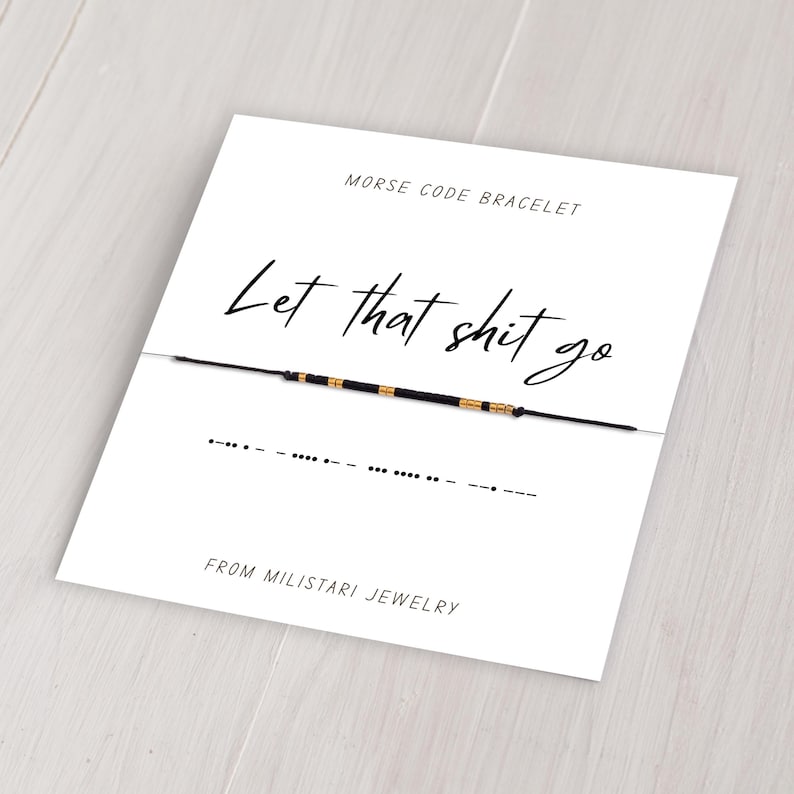 May include: A black string bracelet with gold beads spelling out "Let that shit go" in Morse code. The bracelet is displayed on a white card with the text "Morse Code Bracelet" and "From Miliatari Jewelry".
