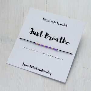 May include: A purple and gold beaded bracelet with the phrase "Just Breathe" and the morse code for the phrase on a white card. The card also says "Morse code bracelet" and "from Milistarjewelry".