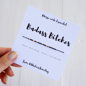 May include: A black string bracelet with gold beads spelling out "Badass Bitches" in Morse code. The bracelet is on a white card with the text "Morse code bracelet" and "from Militstarjewelry".