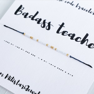 May include: A white card with black text that reads "Badass teacher" and "from Militarijewel". A blue and white beaded bracelet with gold beads is on the card. The bracelet is a code bracelet.