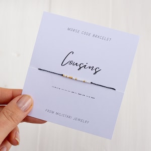 May include: A white card with black text that reads "Morse Code Bracelet" and "Cousins" in cursive. A black string bracelet with white and gold beads is displayed on the card. The Morse code for "Cousins" is shown below the bracelet. The card also includes the text "From Milistasi Jewelry".