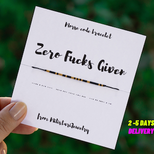 Peut inclure: Un bracelet en perles noires et dorées avec la phrase "Zero Fucks Given" écrite en noir sur une carte blanche. Le bracelet est fait de perles noires et dorées et est attaché avec une ficelle noire. La carte comprend également le texte "Bracelet en code morse" et "de Milistarjewelry".