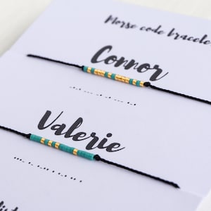 May include: Two black string bracelets with turquoise and gold beads spelling out the names "Connor" and "Valerie" in Morse code. The bracelets are displayed on a white card with the words "Morse code bracelet" written on it.