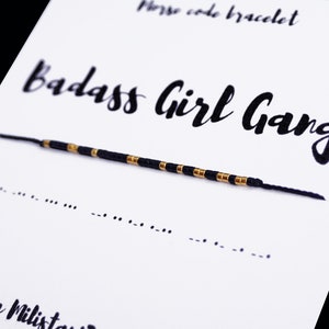 May include: A black and gold friendship bracelet with the words "Badass Girl Gang" written in black cursive on a white card. The bracelet is made of small black and gold beads and is tied with a black string. The card also includes the text "Morse code bracelet" and "from Militant"
