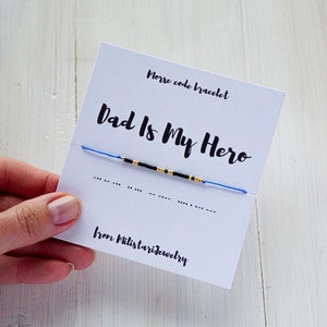 Peut inclure: Un bracelet en perles bleu et noir avec des accents dorés. Le bracelet est sur une carte blanche avec le texte "Dad Is My Hero" et "Morse code bracelet" en noir. La carte comprend également le texte "from Milistarjewelry" et le code Morse de la phrase.