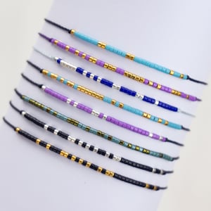 May include: A set of eight colorful beaded bracelets with a variety of colors including turquoise, purple, blue, black, and gold. The bracelets are made with small, square beads and are strung on thin, black cords.