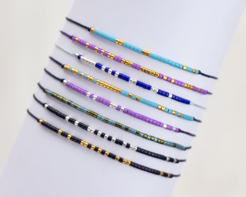 May include: A set of eight colorful beaded bracelets with a variety of colors including turquoise, purple, blue, black, and gold. The bracelets are made with small, square beads and are strung on thin, black cords.