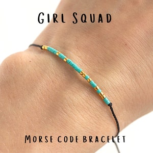 May include: A black string bracelet with turquoise and gold beads spelling out "GIRL SQUAD" in Morse code. The bracelet is on a person's wrist. Morse Code Bracelet.