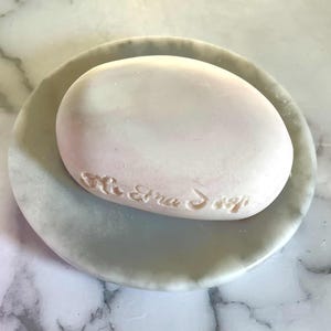 May include: A smooth, oval-shaped light pink bar of soap with embossed text resting on a round, grey and white marbled stone dish. The soap has a soft, matte finish and the dish features natural veining patterns throughout its surface.