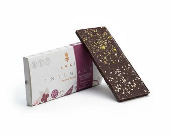 May include: A rectangular dark chocolate bar topped with crushed green pistachios and white salt crystals, leaning against its white and purple paper packaging. The packaging features the text AWKI INTIMACY and floral illustrations.