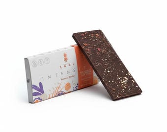 May include: A rectangular dark chocolate bar topped with small nut and fruit pieces, leaning against its white and orange paper packaging. The packaging features the text AWKI INTENSITY Ancestral Wisdom and stylized floral illustrations.
