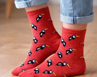May include: A pair of red crew socks featuring a repeating pattern of small toucans with black bodies, white chests, and yellow beaks. The socks have a ribbed cuff at the top and are worn with blue denim jeans that are cuffed at the bottom hem.
