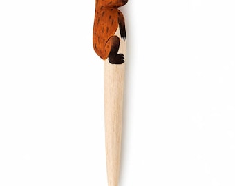 May include: A wooden ballpoint pen featuring a hand-painted cap shaped like a brown capybara. The capybara figure has dark brown markings, a light tan belly, and black feet. The lower barrel of the pen is made of light-colored natural wood.