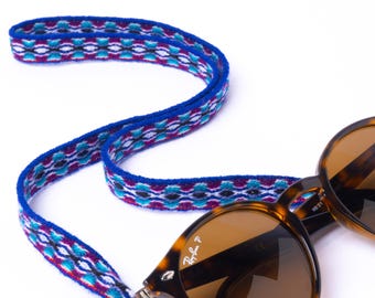 May include: A pair of tortoiseshell sunglasses with brown lenses and a blue, purple, and white patterned strap. The strap is connected to the sunglasses and is designed to be worn around the neck.