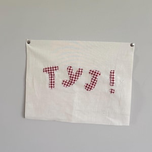 May include: A white fabric banner with the Japanese characters "yuj!" appliqued in a red and white gingham pattern. The banner is hung on a gray wall with two silver tacks.